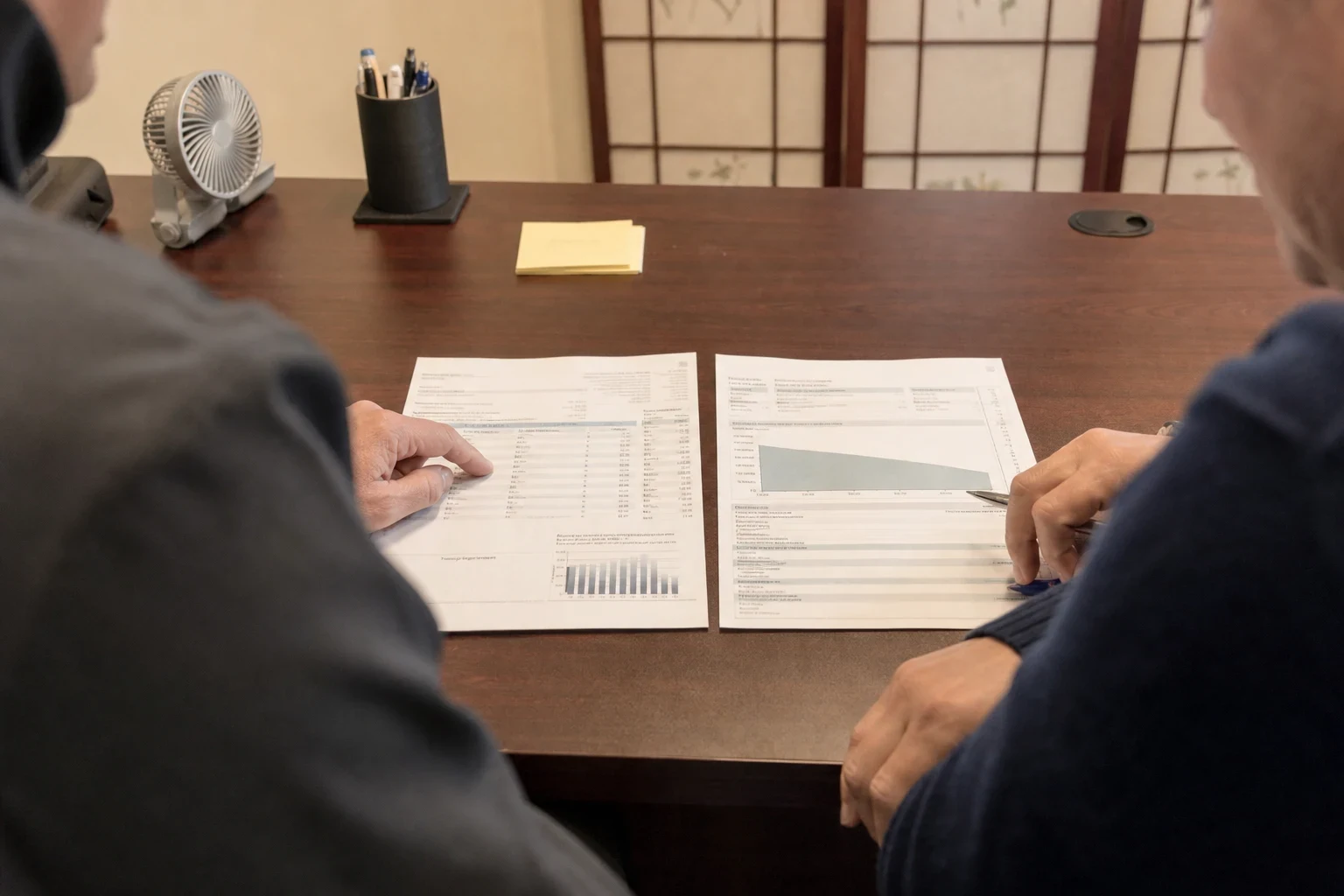 Two people at a wooden office desk reviewing a mortgage statement and a coverage chart laid side by side, one pointing at figures while the other holds a pen