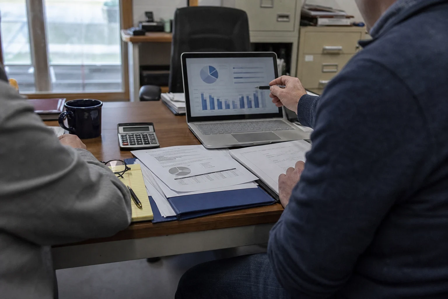 Two people at a wooden office desk reviewing charts on a laptop and printed documents, with a calculator, coffee mug, legal pad and filing cabinets nearby